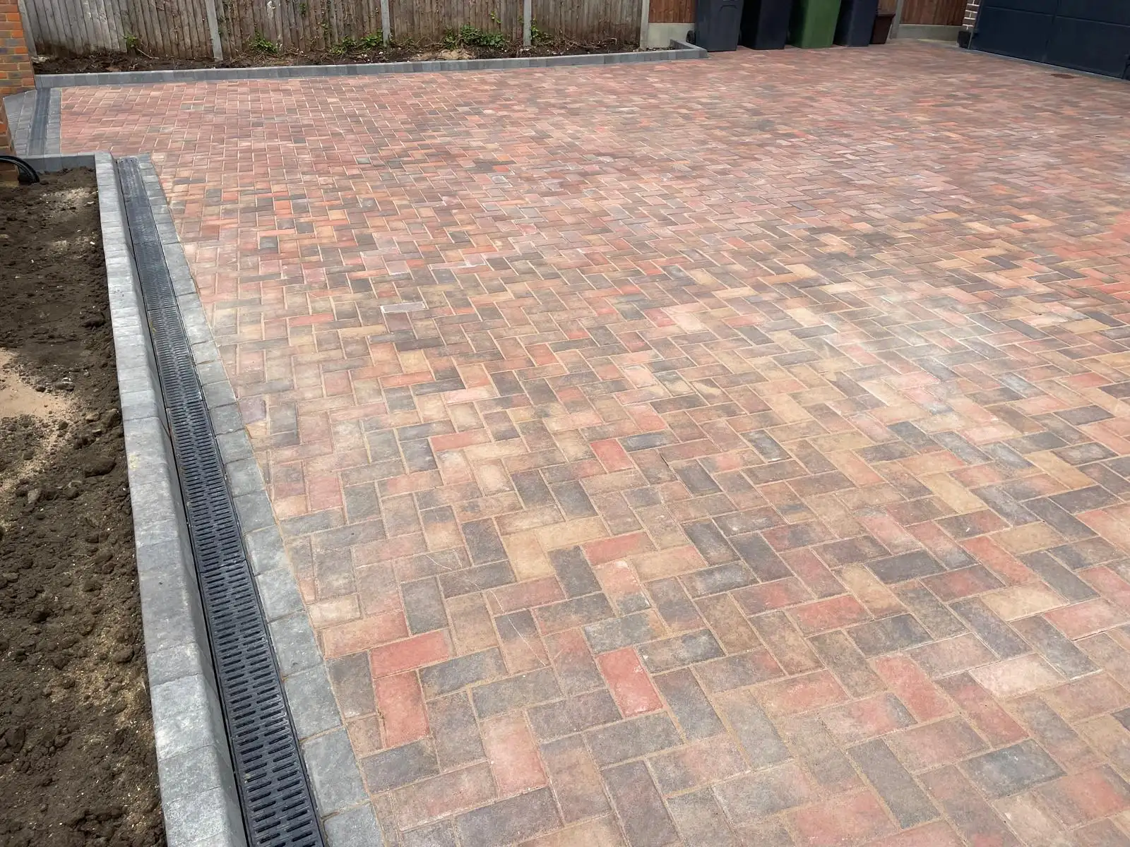 A properly restrained driveway edge with a charcoal kerb course and a linear channel drain set flush, with herringbone paving running up to it.