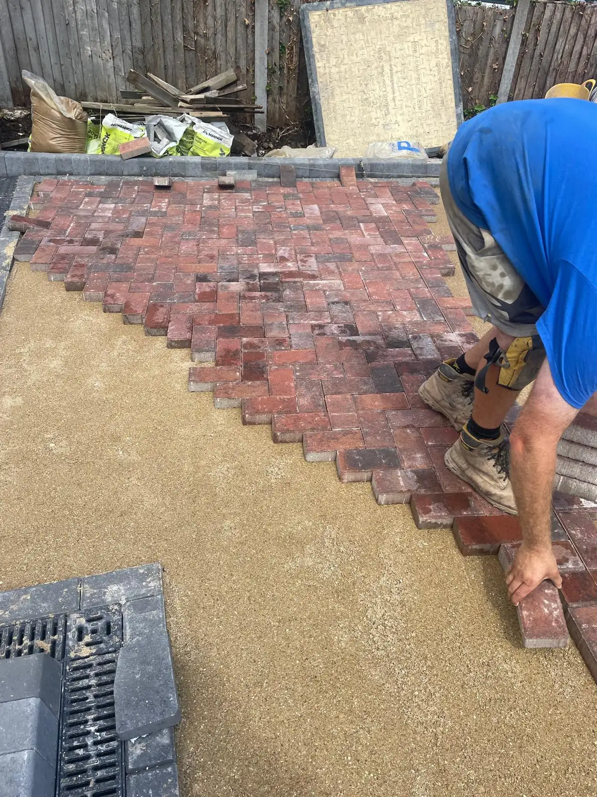 Blocks being laid by hand in a 45 degree herringbone pattern onto a screeded laying course, with the charcoal edging course already bedded and a linear drainage channel in place.